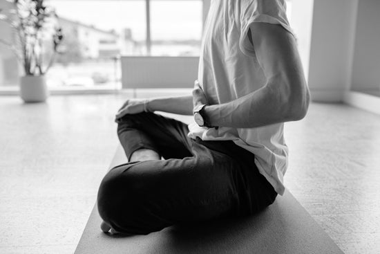 Man sitting cross-legged on a yoga mat on the floor holding his hand to his belly as he practices relaxing breathing exercises.