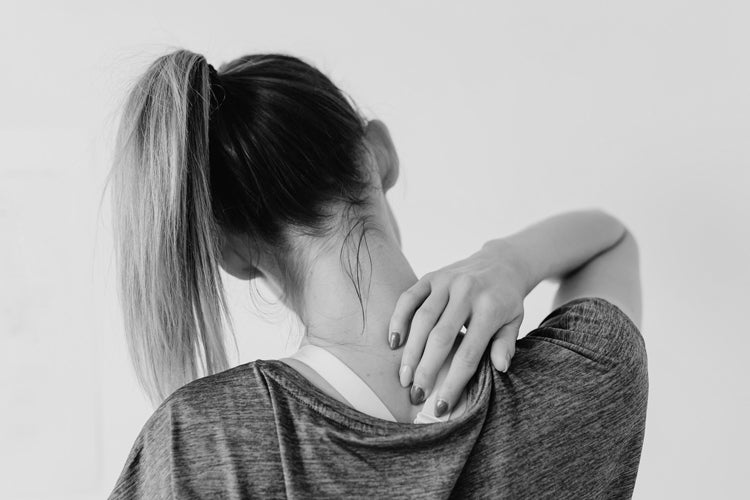 Woman facing away from the viewer reaching back to press her fingers into her aching shoulder and neck.